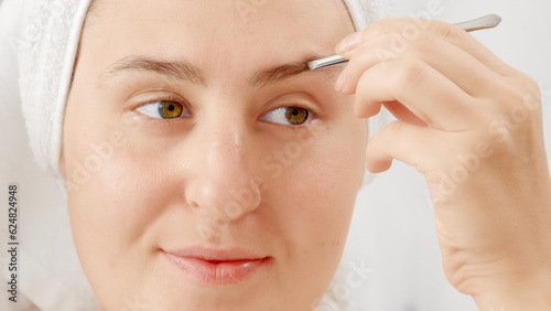 Young woman correcting shape of her yeybrows and plucking them in bathroom. Concept of beautiful female, makeup at home, skin care and domestic beauty industry.