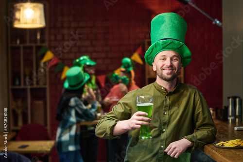 Happy young man with glass of green beer standing by bar counter and looking at camera while celebrating St Patrick day with his friends