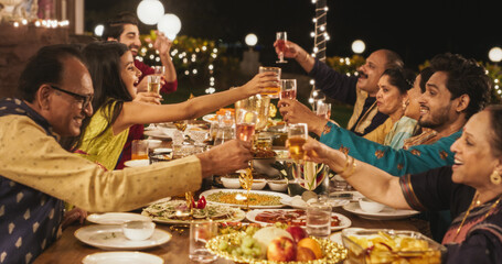 Big Family Celebrating Diwali: Indian Family in Traditional Clothes Gathered Together on a Dinner Table in a Backyard Garden Full of Lights, Making a Toast for Happiness on a Hindu Holiday