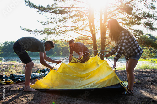 A company of friends guys and girls set up a tent on the sandy bank of the river in summer. Hiking, outdoor recreation in the forest. Sunny day.