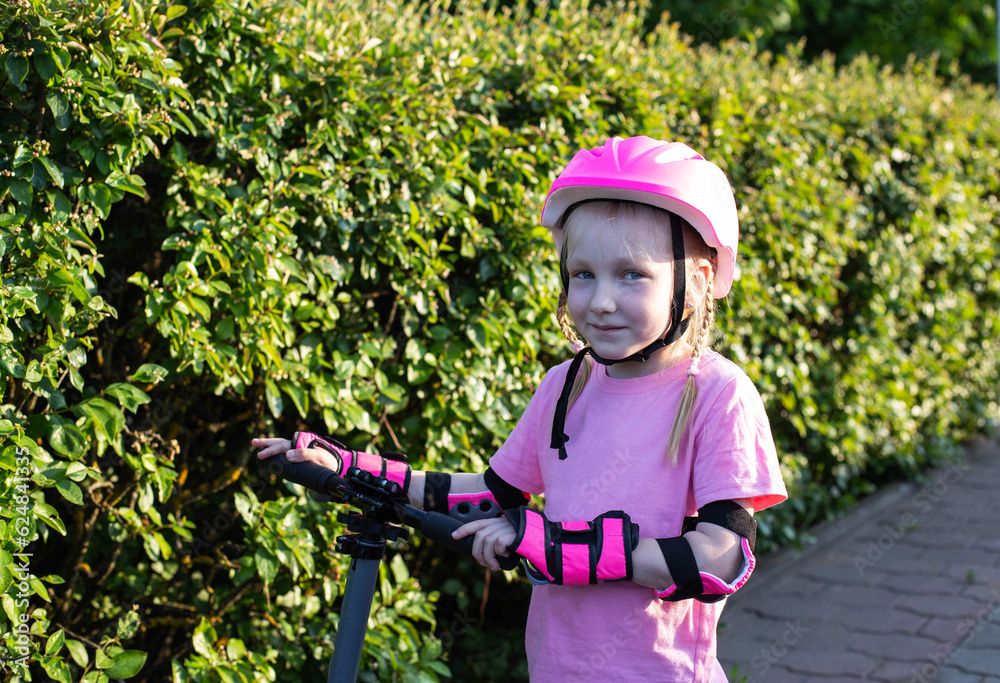 A beautiful girl of 7 years old in a pink helmet and protective sleeves stands against the background of green bushes with a scooter. Safe scooter riding.