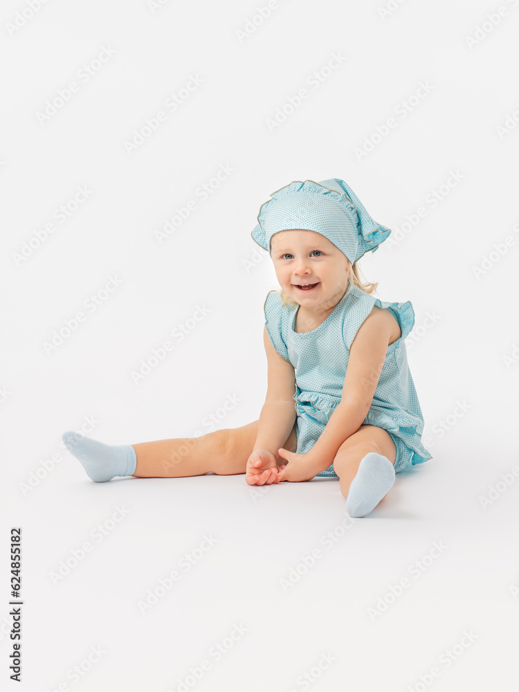 A cheerful blonde toddler girl of 2 years old in a blue dress and a kerchief sits with her feet on the floor, laughs, smiles and looks at the camera on a white background. Photo A cheerful blonde toddler girl of 2 years old in a blue dress and a kerchief sits with her feet on the floor, laughs, smiles and looks at the camera on a white background. Photo