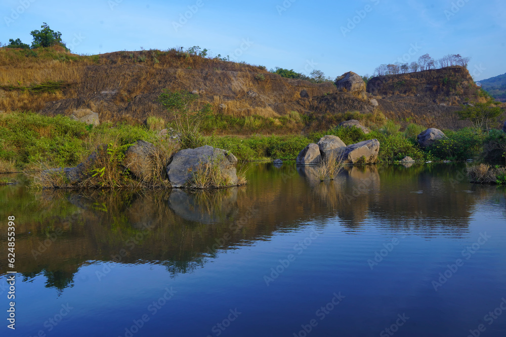 A serene pond reflects the surrounding landscape of lush grass, scattered rocks, and trees in Ranu Manduro, Mojokerto, East Java, Indonesia.