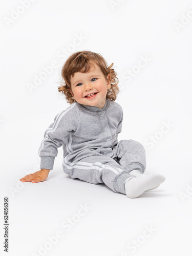 The toddler is 2 years old. A joyful boy is sitting on the floor and laughing with his head tilted with curly hair, plump cheeks in a gray jumpsuit and socks on a white background.