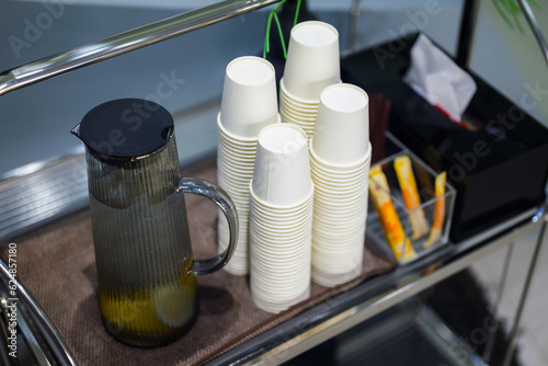 Free drinking water for self service in coffee shop, stacks of single-use cups and cold water jug on the rack wheels