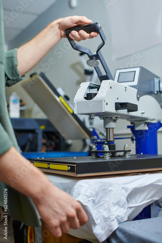 Cropped view of young artisan working with screen printing machine and clothes in blurred print studio at background, customer-focused small business concept