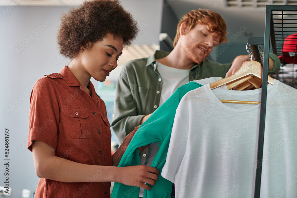 Obraz premium Smiling young african american craftswoman looking at clothes on hangers near blurred redhead colleague and standing together in print studio, young small business owners concept