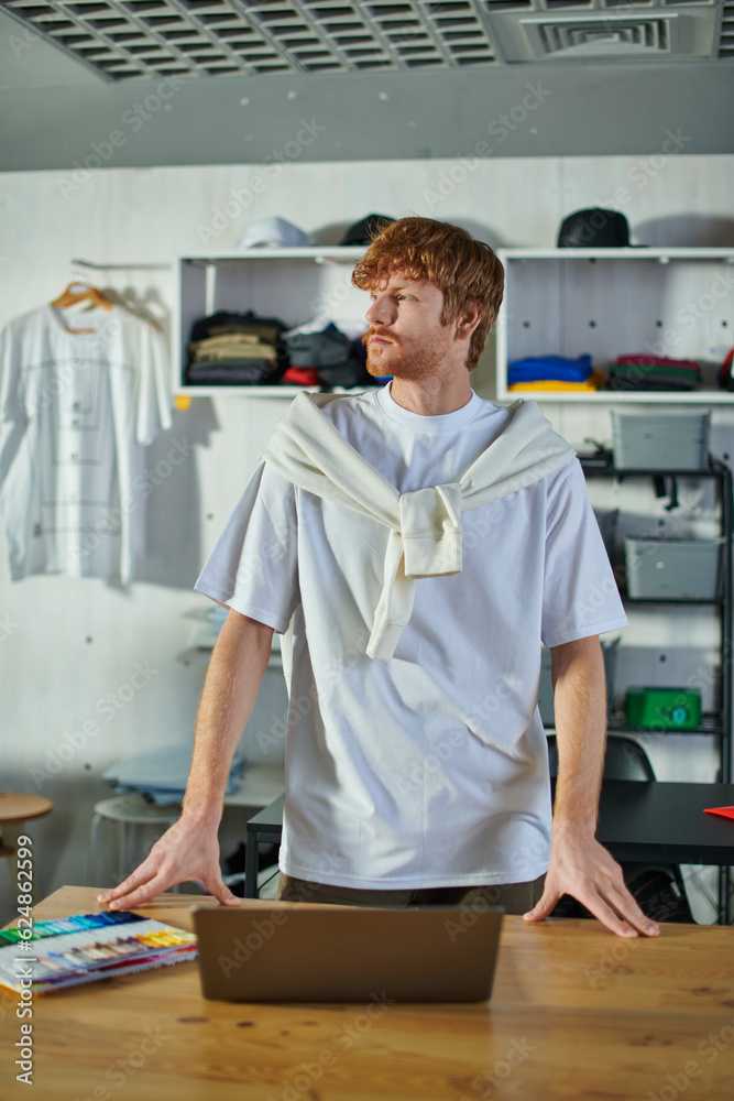 Young redhead craftsman in casual clothes looking away while standing near laptop and cloth samples on table in blurred print studio, self-made success concept