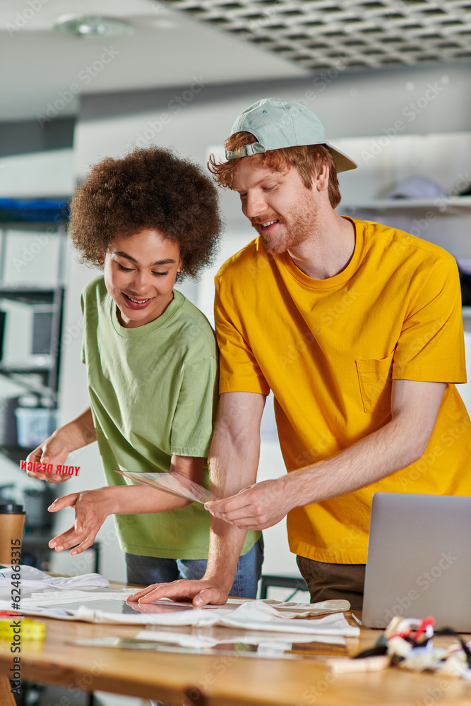 Smiling craftsman holding printing layer near african american colleague, clothes and laptop on table while working in blurred print studio at background, start-up innovation concept