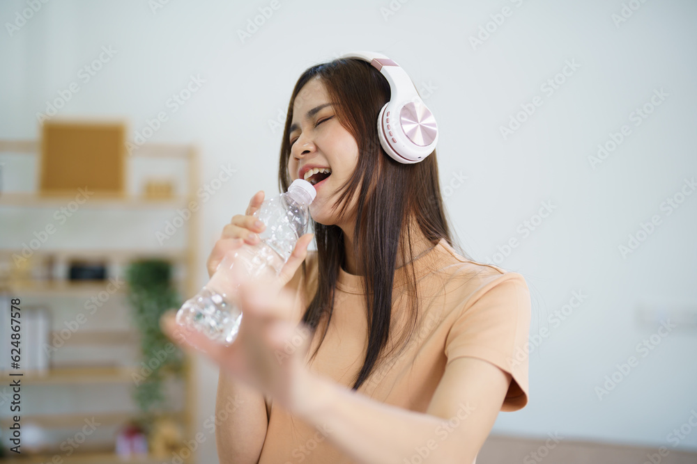 Happy Young Asian woman singing alone with bottle like microphone and wearing headphone love music happy relax in living room Happy Young Asian woman singing alone with bottle like microphone and wearing headphone love music happy relax in living room