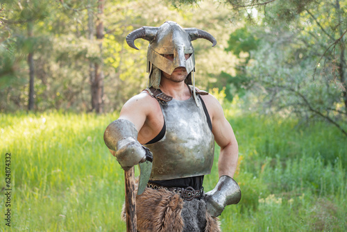 Portrait of a warrior in a horned helmet, a steel breastplate with a two-handed ax in his hands, posing against the backdrop of the forest. 