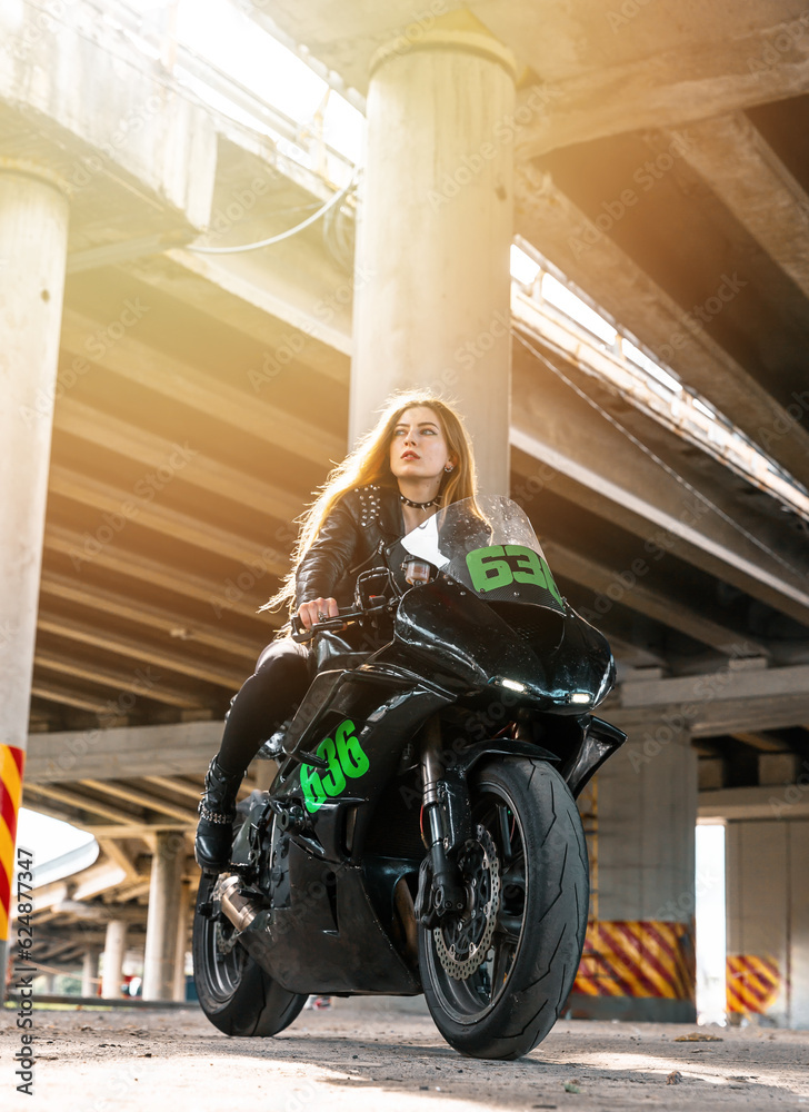 Woman in black leather jacket and jeans pants on outdoors parking with stylish sports motorcycle at sunset. Rider under highway