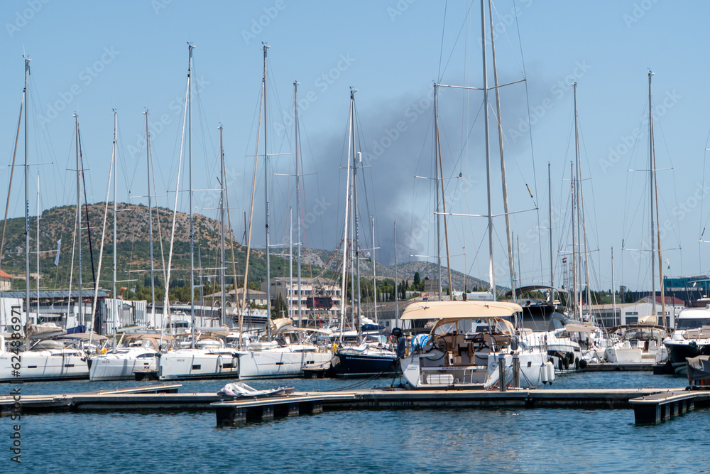 Obraz premium Waldbrand bei Sibenik gesehen durch einen Hafen mit Segelbooten im Vordergrund