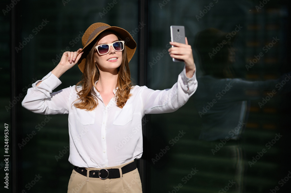 Young happy woman in summer clothes, sunglasses, straw hat makes a selfie portrait, blogs near the business center Young happy woman in summer clothes, sunglasses, straw hat makes a selfie portrait, blogs near the business center