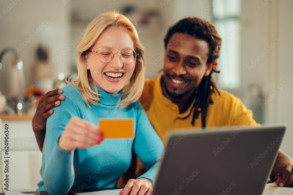 A cheerful interracial couple is sitting at their cozy home and shopping online on retail on the laptop. A cheerful interracial couple is sitting at their cozy home and shopping online on retail on the laptop.