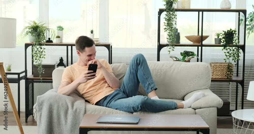 Caucasian man is seated comfortably on a couch, engrossed in his smartphone. With a relaxed posture, he effortlessly balances the device in his hand, his fingers tapping and scrolling with ease. Caucasian man is seated comfortably on a couch, engrossed in his smartphone. With a relaxed posture, he effortlessly balances the device in his hand, his fingers tapping and scrolling with ease.