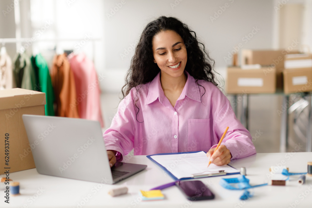 Fashion Entrepreneur Makes Notes Works On Laptop In Clothing Showroom Fashion Entrepreneur Makes Notes Works On Laptop In Clothing Showroom