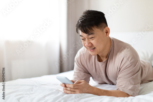 Happy asian man using phone in bed, using mobile app