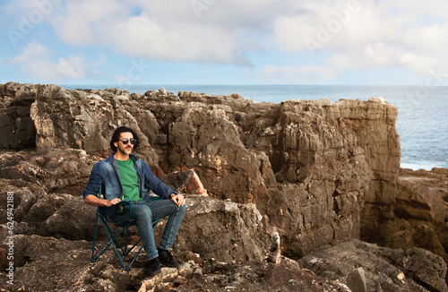Handsome Young Man Relaxing In Camping Chair On Beachrocks Near Ocean