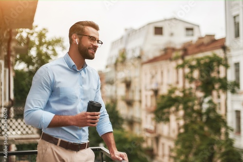 Good news. Young and handsome bearded man in eyeglasses and headphones holding cup of coffee and talking with friend while standing at the office balcony