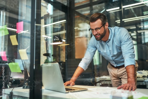 Great results Young bearded businessman in eyeglasses and formal wear looking at laptop and smiling while standing in the modern office