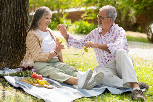 Happy senior european man feeding grapes to lady, enjoy romantic date, lunch in park, have fun © Prostock-studio