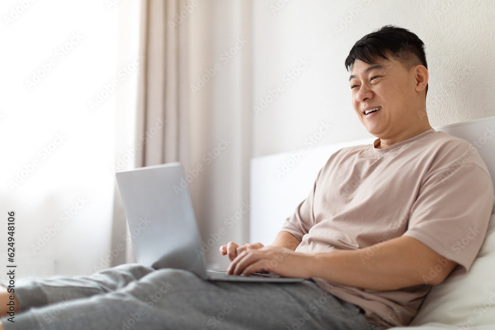 Happy mature asian man using laptop in bed, copy space Happy mature asian man using laptop in bed, copy space