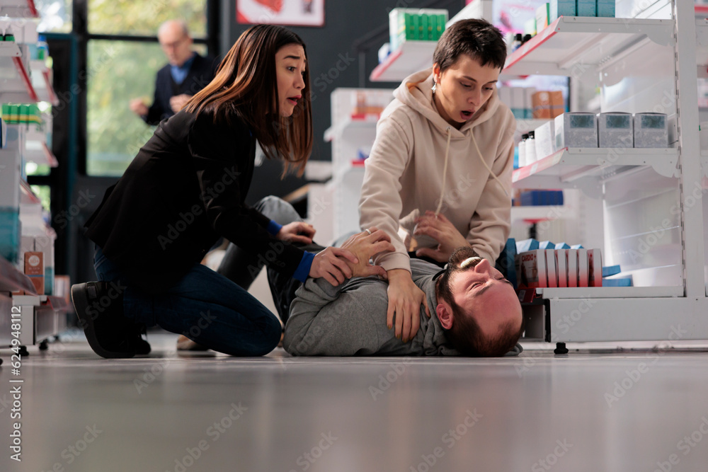 Frightened women rescuing man suffering from epilepsy attack, holding unconscious person and waiting for help in drugstore. Pharmacy asian and caucasian clients noticing epileptic seizure accident