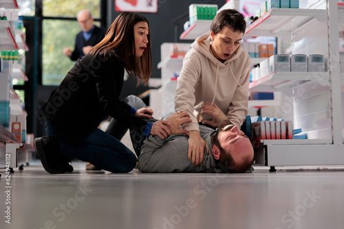 Frightened women rescuing man suffering from epilepsy attack, holding unconscious person and waiting for help in drugstore. Pharmacy asian and caucasian clients noticing epileptic seizure accident photo