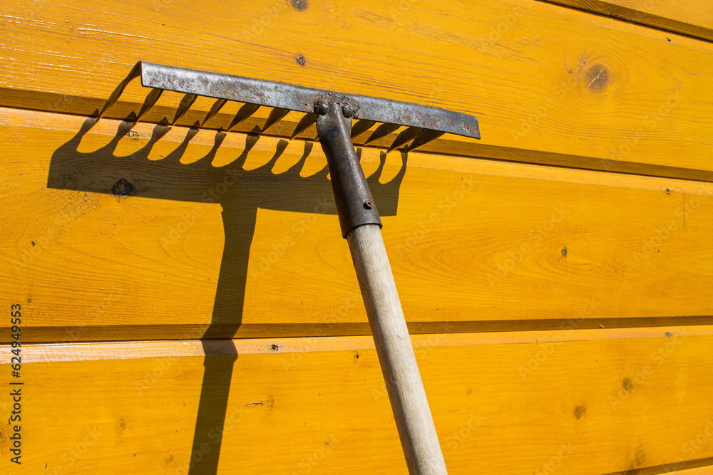 An old rusty rake is leaning against a yellow wooden wall and a contrasting shadow and copy space.  Concept - gardening and countryside