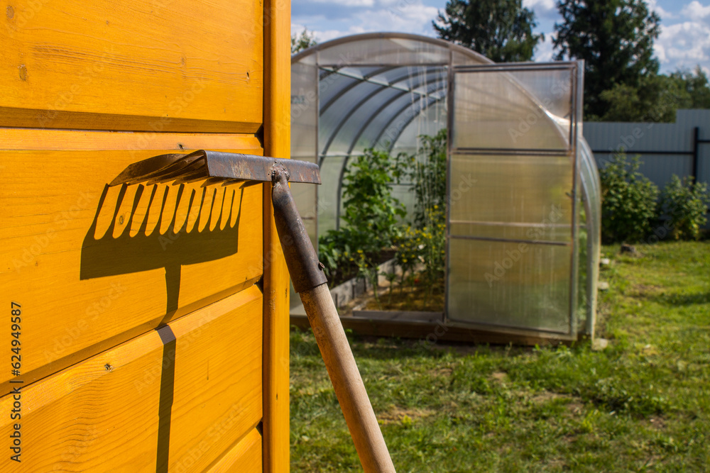 An old rusty metal rake near a yellow wooden wall and a bright contrasting shadow on a sunny day. The concept is gardening and agriculture