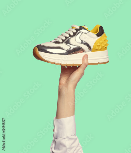 Konstfotografi A woman's hand holding a fancy sneaker with a chunky insole, shiny silver inserts and a yellow counter with spikes, isolated on a minty green background