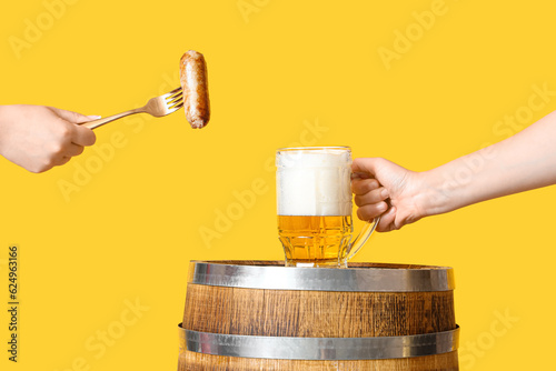 Female hands with sausage and mug of cold beer on yellow background