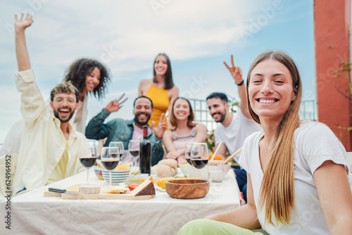 Group of happy people taking a selfie on a rooftop lunch party celebration, laughing together. Smiling woman looking at camera and enjoying a meal with her best friends on a friendly diner meeting