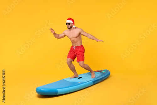Happy man in Santa hat balancing on SUP board against orange background © New Africa