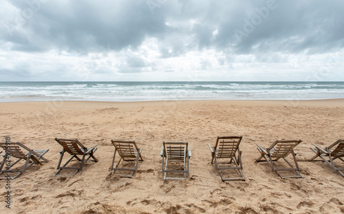 Leinwand Poster Espreguiçadeiras de madeira vazias na areia da praia deserta de frente para o mar em dia nublado