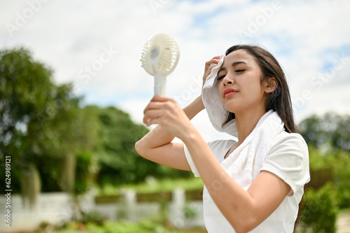 An exhausted and sweaty Asian woman using a portable handy fan, feeling hot and tired