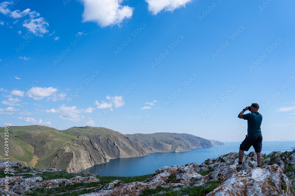 person on the top of mountain. A young man is standing on a rock and looking at the sea through binoculars. Lake in summer. A tourist is resting on the shore and looking through binoculars. 