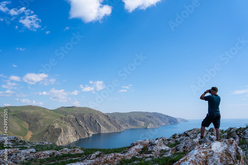 person on the top of mountain. A young man is standing on a rock and looking at the sea through binoculars. Lake in summer. A tourist is resting on the shore and looking through binoculars.  photo