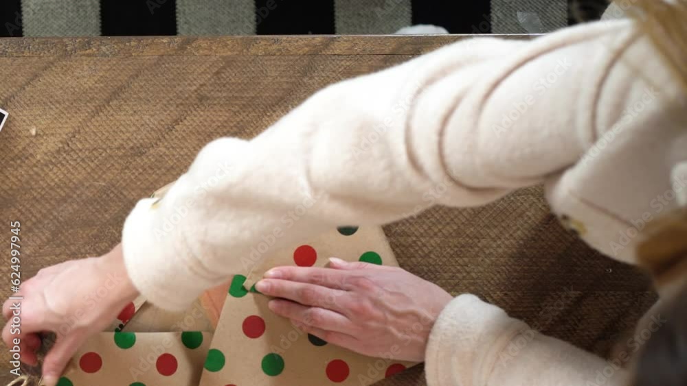 A young woman wearing a fleece coat struggles to wrap a holiday Christmas present with decorative polka dot wrapping paper and tape on a wooden table.