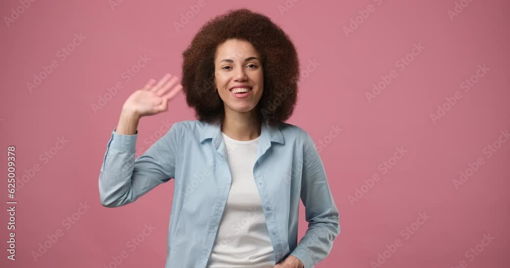 Happy friendly young african american woman waves palm in hello gesture welcomes someone with hospitable expression expresses positive emotions standing over pink background Happy friendly young african american woman waves palm in hello gesture welcomes someone with hospitable expression expresses positive emotions standing over pink background