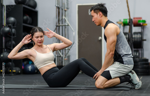 Asian strong young male muscular fitness trainer helping female athlete model in sexy sport bra and legging lifting laying down sit up on floor exercise training in gym.