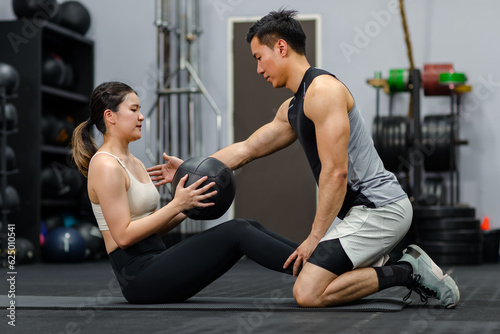 Asian strong young male muscular fitness trainer helping female athlete model in sexy sport bra and legging lifting working out with heavy leather ball workout on floor exercise training in gym