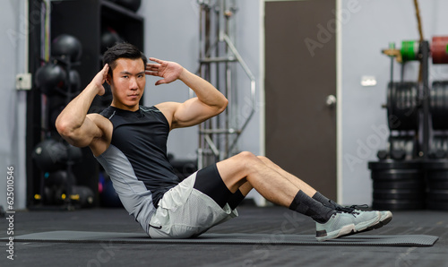 Millennial Asian strong young fit male muscular fitness model in sportswear sleeveless shirt and sporty shorts laying down sit up looking at camera on mat floor working out exercise training in gym
