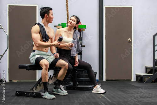 Asian strong young male muscular shirtless fitness model in sporty shorts and female athlete in sport bra sitting smiling taking break holding water bottle talking together in gym