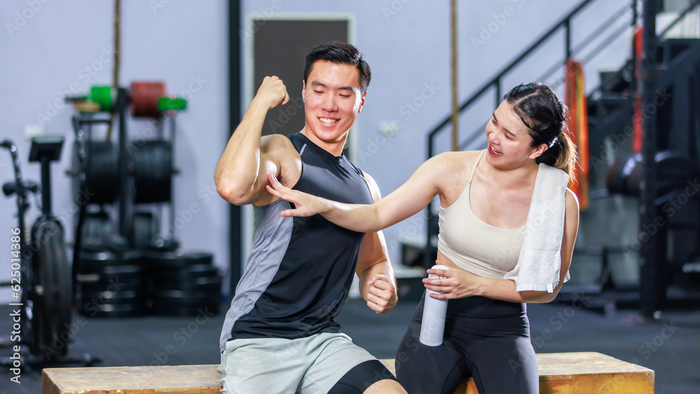 Asian strong young fit male muscular fitness athlete model showing off bicep muscle to female friend sitting take break drink water wipe sweat on wood box smiling hold thumb up complimenting in gym Asian strong young fit male muscular fitness athlete model showing off bicep muscle to female friend sitting take break drink water wipe sweat on wood box smiling hold thumb up complimenting in gym