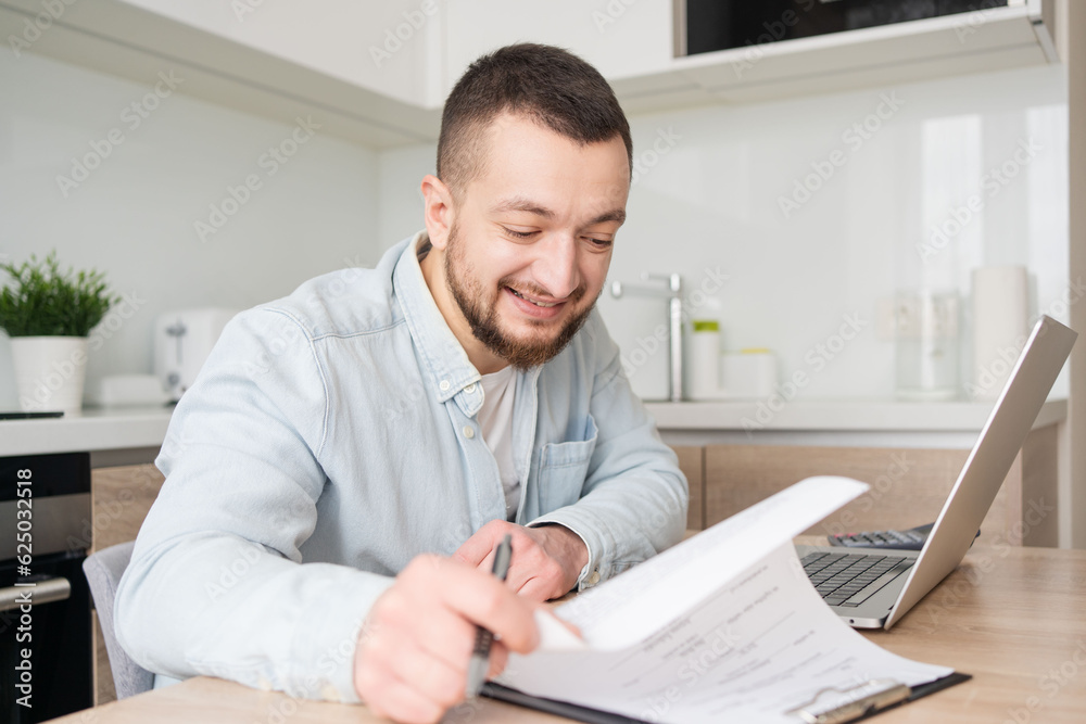 young man watching educational webinar lecture on computer, writing notes on paper at home office. Serious businessman analysing electronic marketing documents indoors