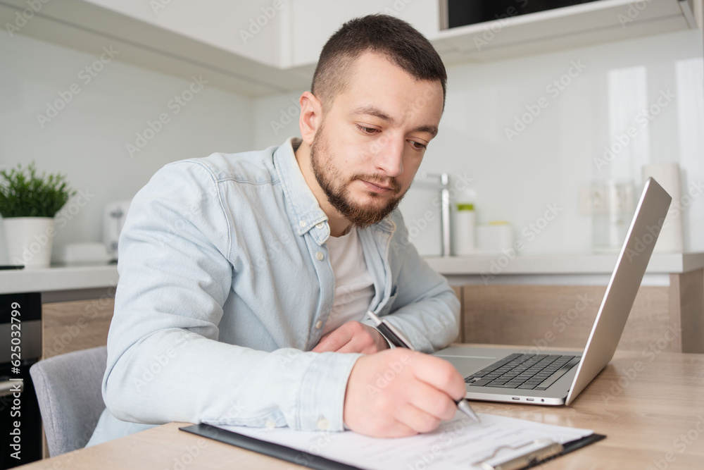 young man watching educational webinar lecture on computer, writing notes on paper at home office. Serious businessman analysing electronic marketing documents indoors young man watching educational webinar lecture on computer, writing notes on paper at home office. Serious businessman analysing electronic marketing documents indoors