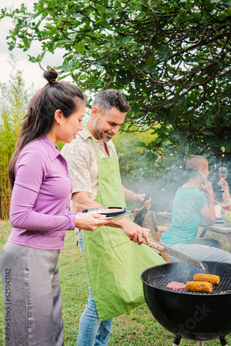 Vertical portrait. Middle adult happy couple cooking meat on a barbecue bbq party with their friends at home backyard. Husband and wife grilling food together on barbeque weekend friendly celebration