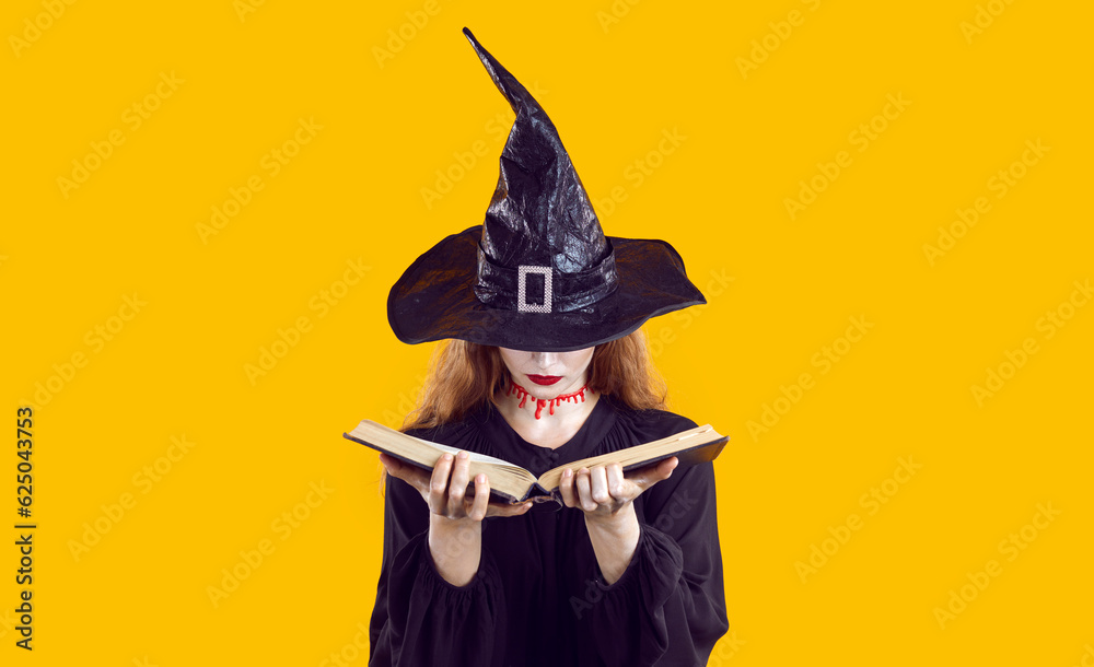 Young girl in carnival costume of a witch in hat standing in the studio isolated on orange background. Woman holding a book in her hands reading a spell and conjuring. Halloween and magic concept.